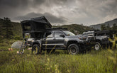 Two Toyota Tacoma overland builds with deployed rooftop tents in a Colorado mountain meadow at dusk